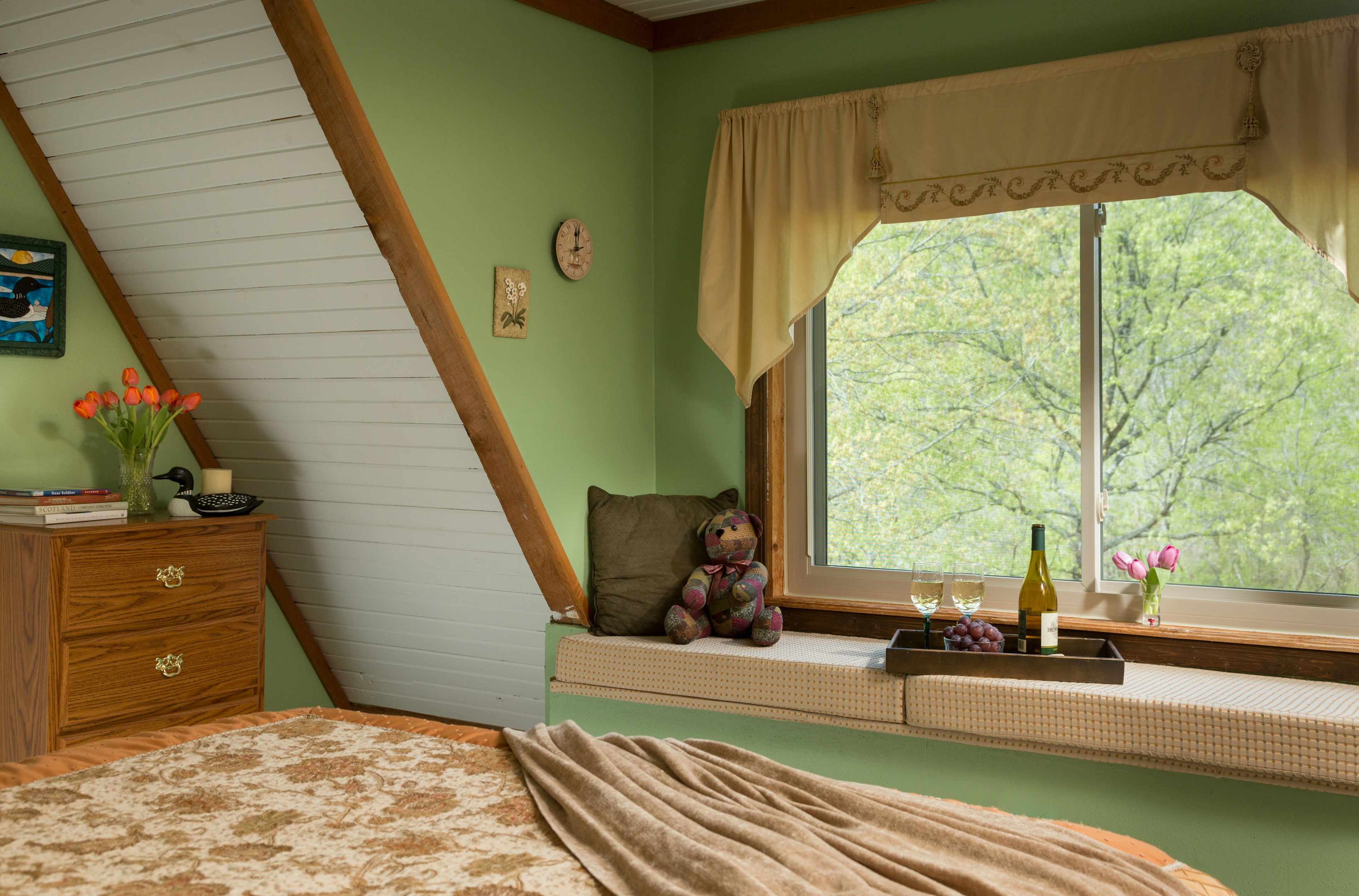 bedroom with part of a bed in the foreground.  On the bed are a quilt and throw blanket.  In front of the bed is a window seat with cushions.  On top of the cushions are a pillow, a stuffed bear, and a tray with a wine bottle and two glasses.  There is a large window above the window seat, overlooking trees.  To the left of the windowseat is a wooden dresser with flowers and books on top.