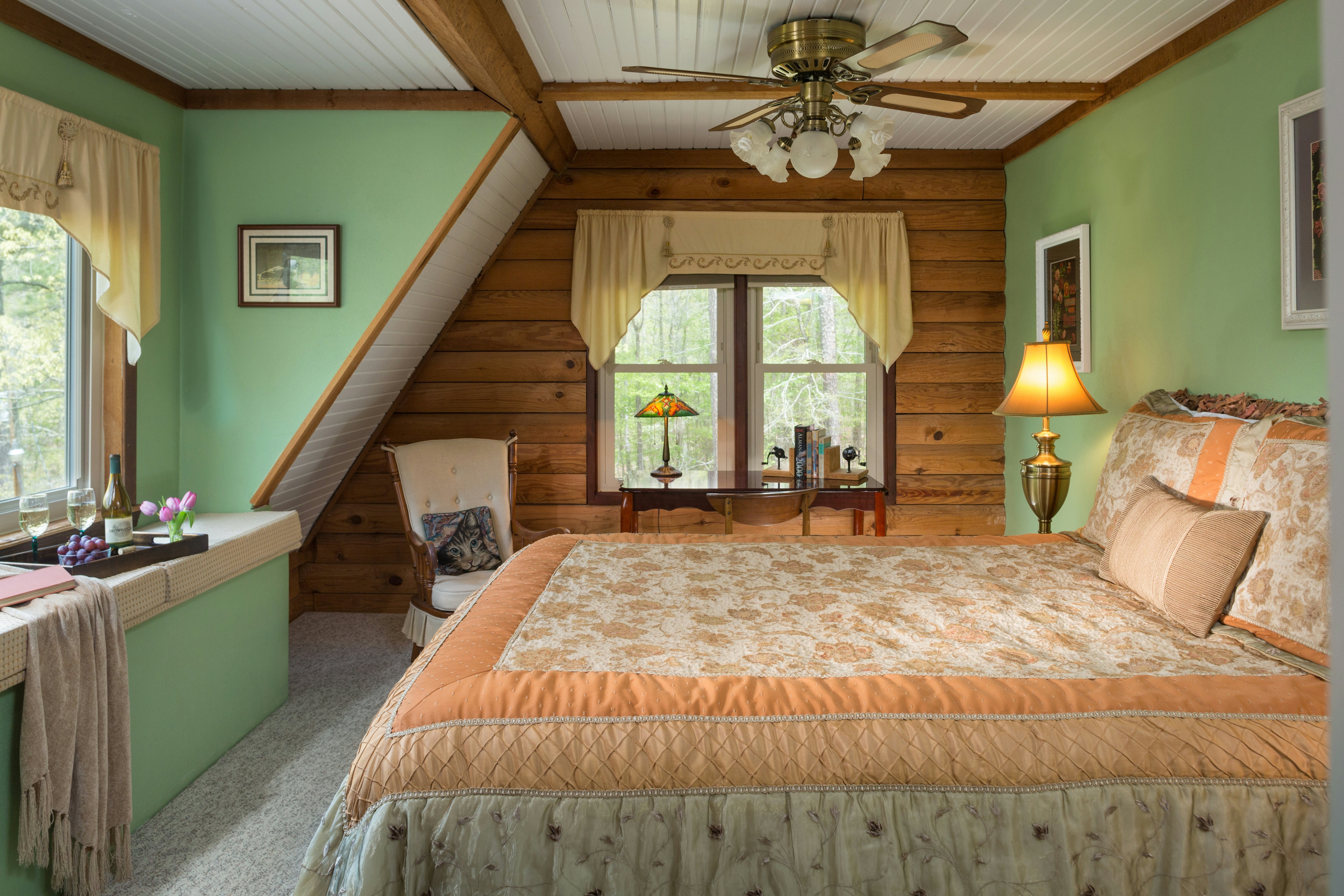 A bedroom with two green walls and one log wall.  A queen-sized bed is in the middle, with a quilt and decorative pillows. on the left is a window seat with cushions.  On top of the cushions is a throw blanket and a tray with a wine bottle and wine glasses.  In the background is a padded rocking chair, and a desk with a table lamp and books on it. The desk is sitting in front of a window that overlooks trees outside.  There is carpet on the floor, and a ceiling fan on the ceiling.