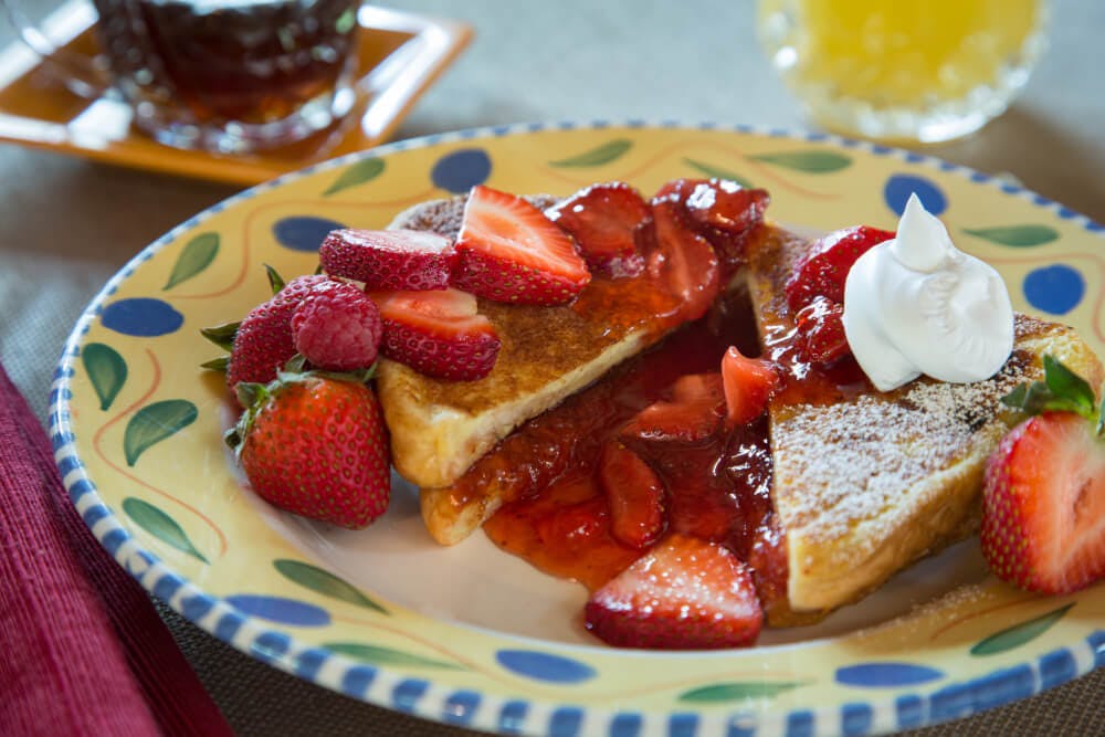 A colorful plate with stuffed French toast dusted with powdered sugar, and topped with sliced strawberries and a dollop of whipped cream.