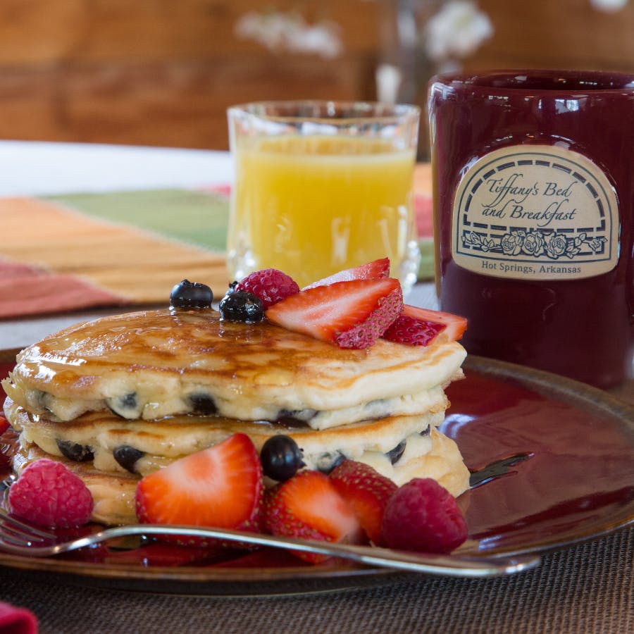 A plate of blueberry pancakes topped with sliced strawberries, with a glass of orange juice and a red coffee mug that says Tiffany’s Bed & Breakfast.