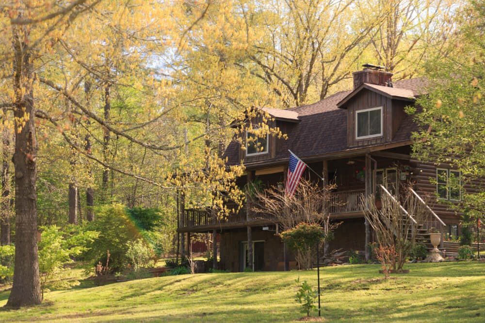 A multi story log home surrounded by grassy grounds, trees, and woods