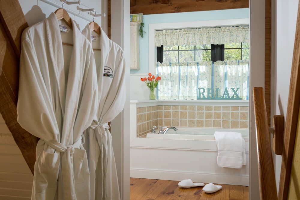 A bathroom with bathrobes hanging on hooks, a jetted bathtub, and a window ledge with a vase of tulips and a sign that says relax.