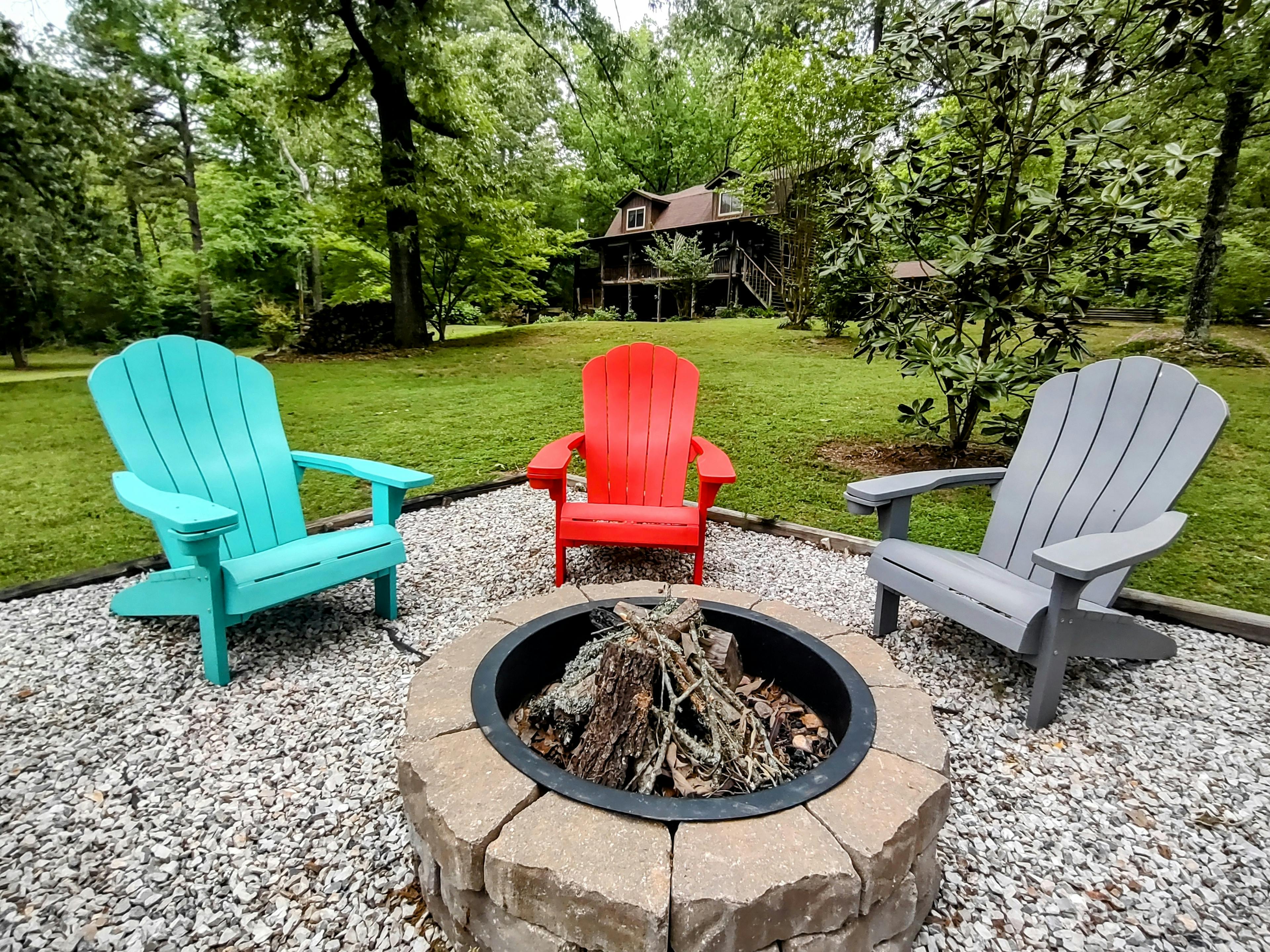 Green, red and gray adirondack chairs around a fire pit in front of a grassy yard with a log house in the background