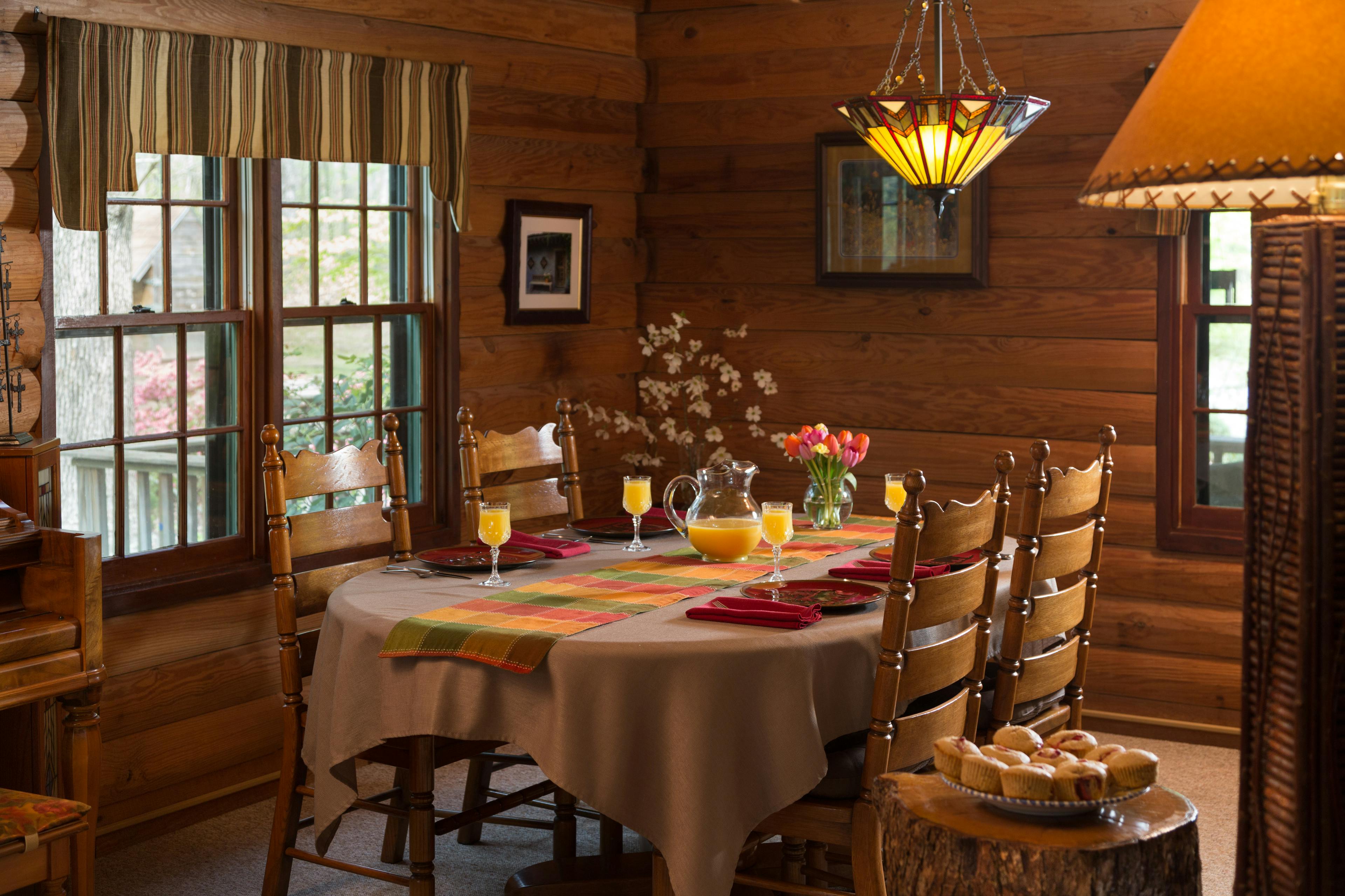 A log dining room with an oval table, brown tablecloth, orange, red and green table runner, red plates and napkins, and glasses full of orange juice, along with wood chairs.