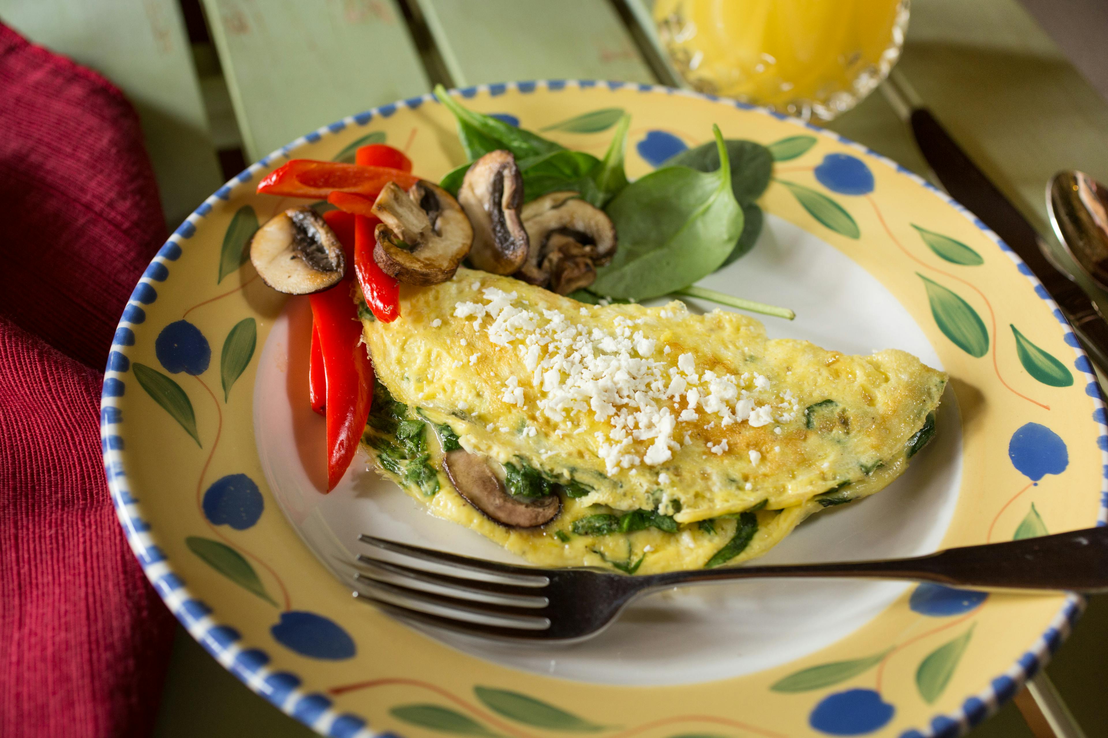 An omelet with sauteed spinach and mushrooms with a side of fresh vegetables on a white plate with a colorful rim