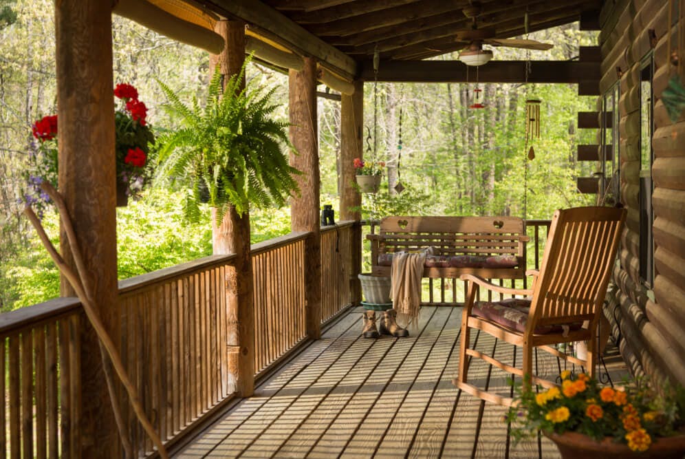A wood porch with wood rocking chairs and a porch swing, with hanging potted flowers, overlooking the grounds and woods.