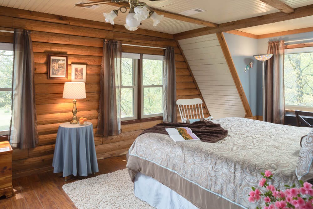 A bedroom with log walls, wood floors with a carpeted area rug, a bed with blue and brown tapestry bedding, a small table with a lamp in between 2 windows, and a rocking chair in the corner.