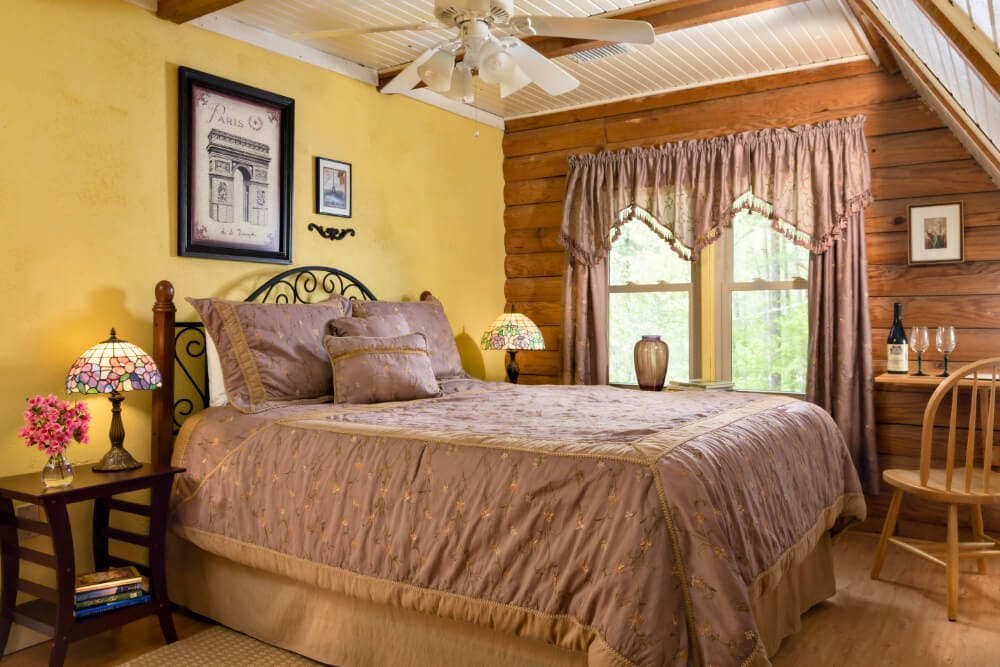 A bedroom with yellow and log walls, wood floors, a bed with a wrought iron headboard, and nightstands and lamps on either side of the bed.