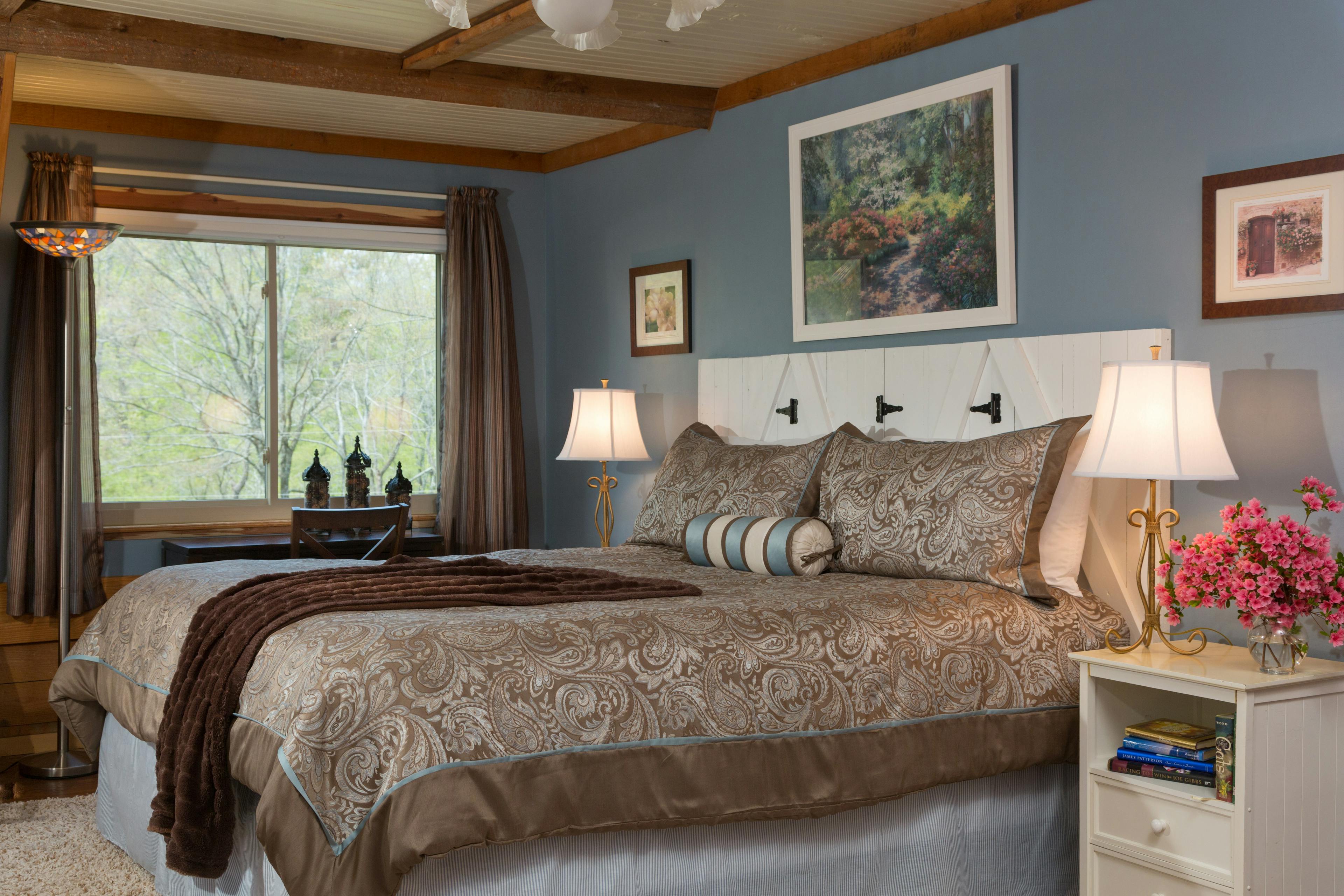king-sized bed with 2 large and one small accent pillows, a brown throw blanket, and a brown & blue duvet cover.  To the right of the bed is a white nightstand wtih books on the shelf, and a table lamp and pink flowers on top.  To the left of the bed is a matching lamp, and a small desk in front of a large window.