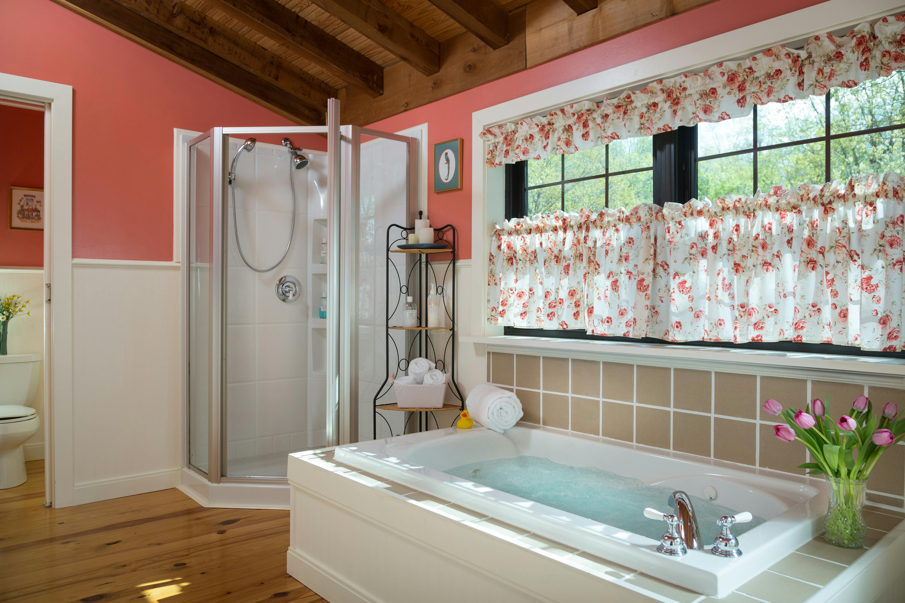 Large bathroom with a stand alone shower with a glass door.  Next to the shower is a small corner shelf with bath accessories.  To the right of the shelf is a jetted tub filled with water, and windows with pink & white curtains.