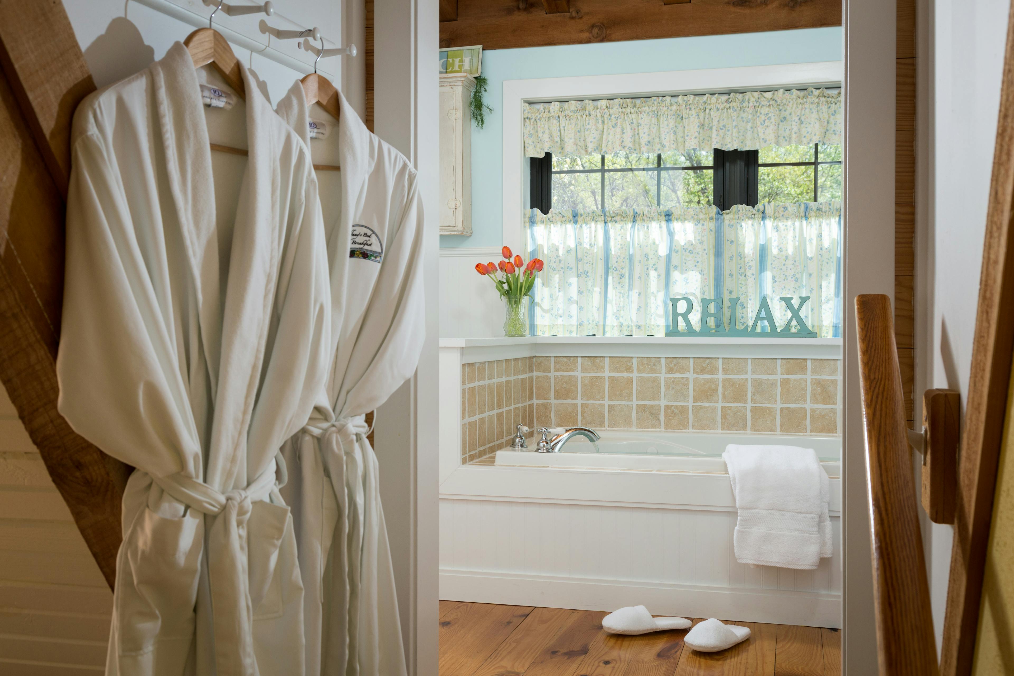 Two white robes hanging on a wall beside stairs leading to a bathroom with a wooden floor.  In the bathroom is a jetted tub with white towels hanging off the edge.  There is a pair of white slippers in front of the tub.  Behind the tub are two windows covered with blue & yellow curtains.