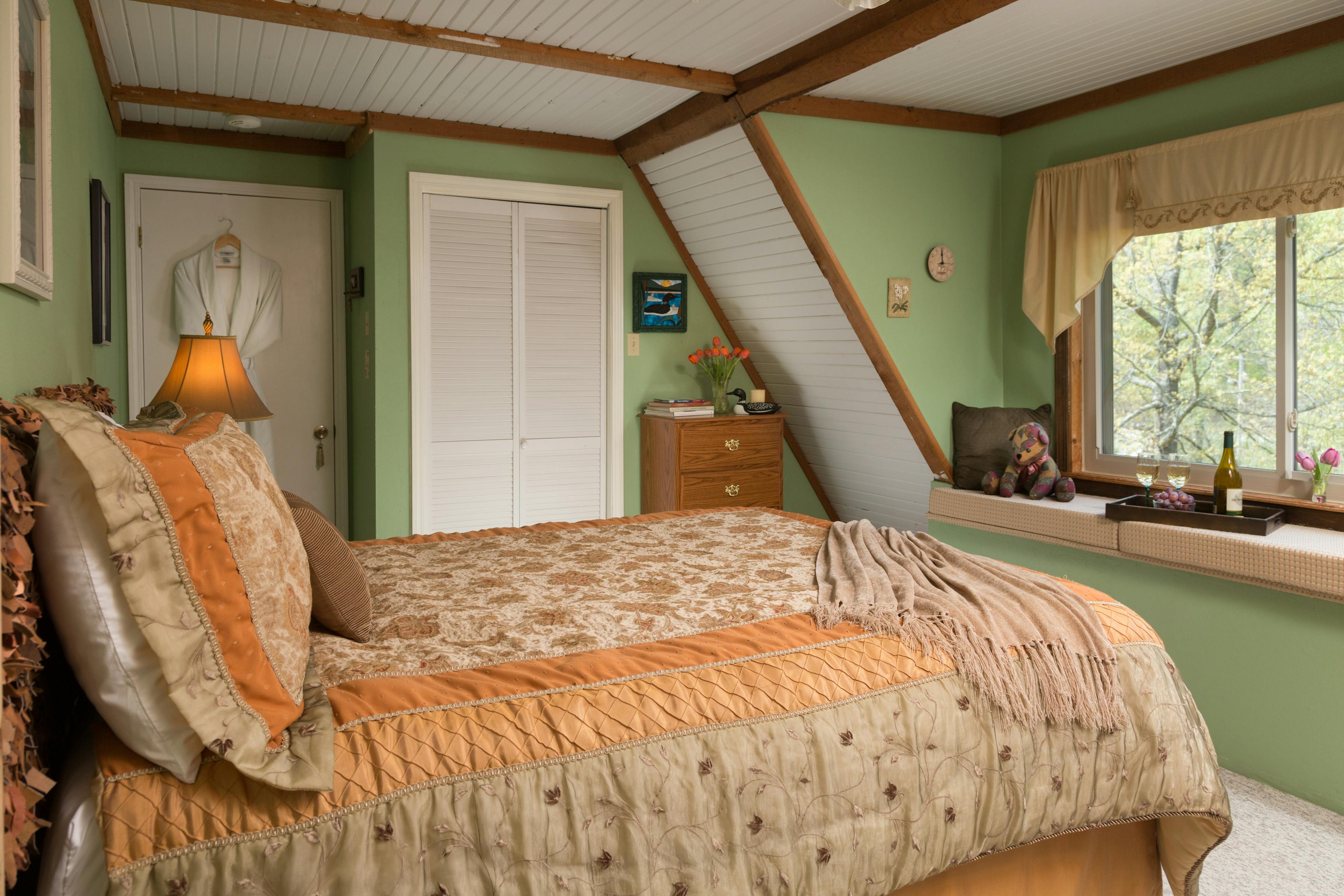 A bedroom with a queen sized bed with rust colored quilt and decorative pillows.  Behind the bed is a white door with a white robe hanging on it.  To the right of that door is a white closet door, and a wooden dresser with books and a vase of flowers on top.  The bed is facing a window seat with cushions and a tray with a bottle of wine and wine glasses, and a decorative pillow.  The window seat is in front of a large window overlooking trees.  The bedroom walls are white and green.  There is carpet on the floor.