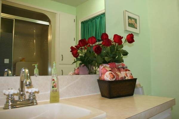 A bathroom with green walls and white trim.  A shower is reflected in the vanity mirror.  On the vanity is a sink, a basket of soaps, and a vase with red roses.
