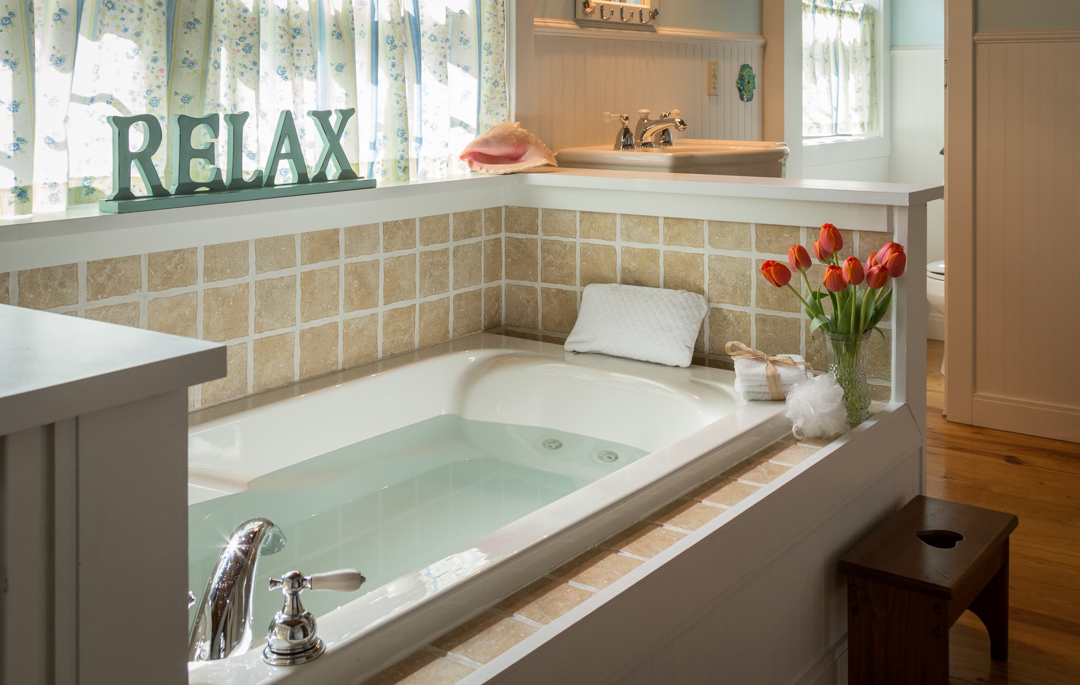 The private bathroom in The Hamilton room at Tiffany's Bed and Breakfast near Hot Springs, AR. The photo features a large, deep jetted soaking tub built into a white-tiled alcove with a 'RELAX' sign and fresh orange tulips. The space includes a pedestal sink, soft natural lighting from curtained windows, and a boutique bed and breakfast atmosphere in Bismarck.