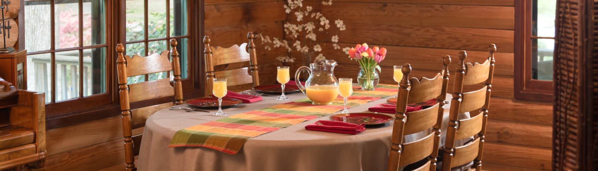 A rustic dining room with a table set for breakfast with a colorful table runner, red plates and napkins, and glasses of orange juice.